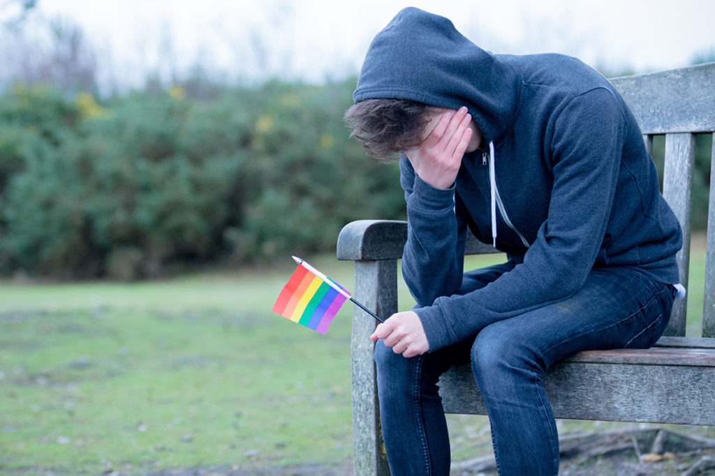 A young man holds a LGBTQ flag and covers his face as he remembers that lgb teens are more likely to binge drink