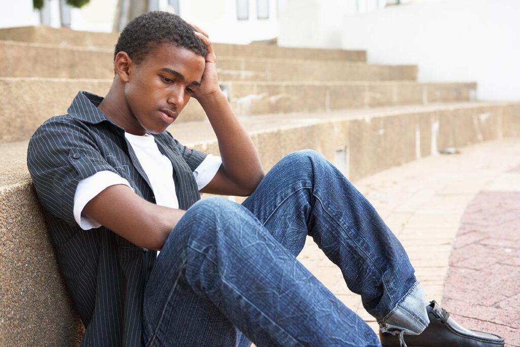 a young person holds their head while sitting on the ground possibly struggling with teen drinking