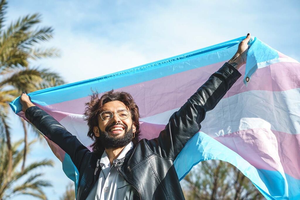 a trans person holds a trans flag up outside next to palm trees to show a transgender drug rehab