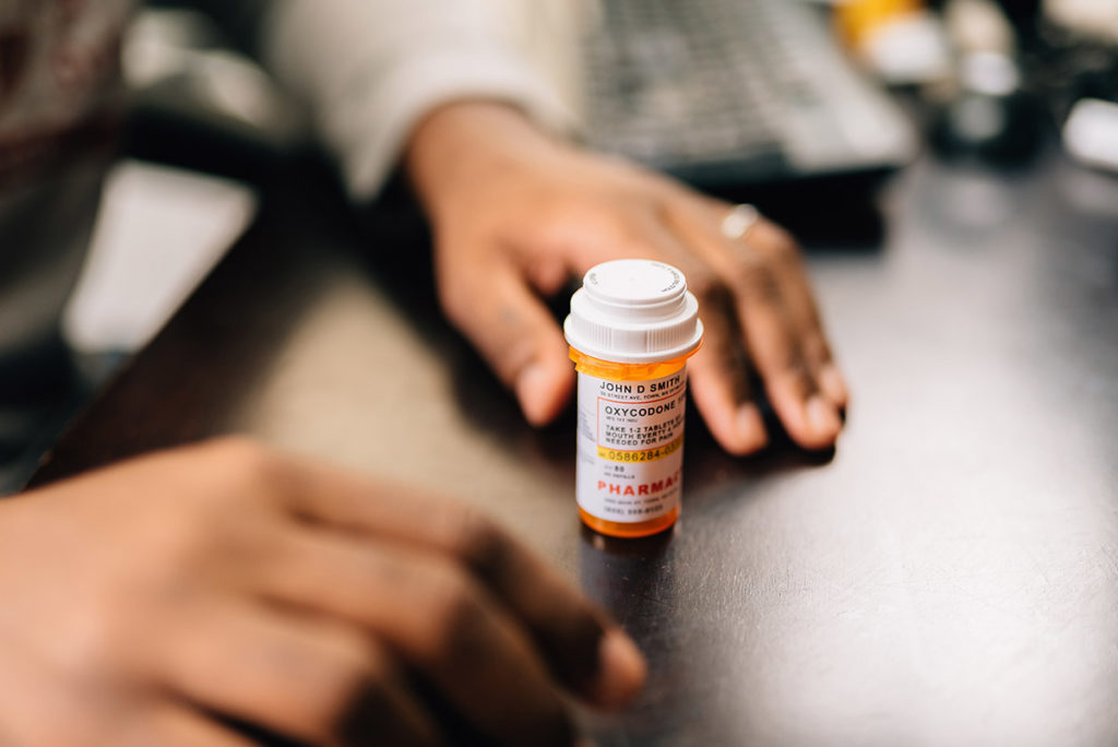 person hesitating to take pill from prescription bottle on desk after reading opioid epidemic statistics