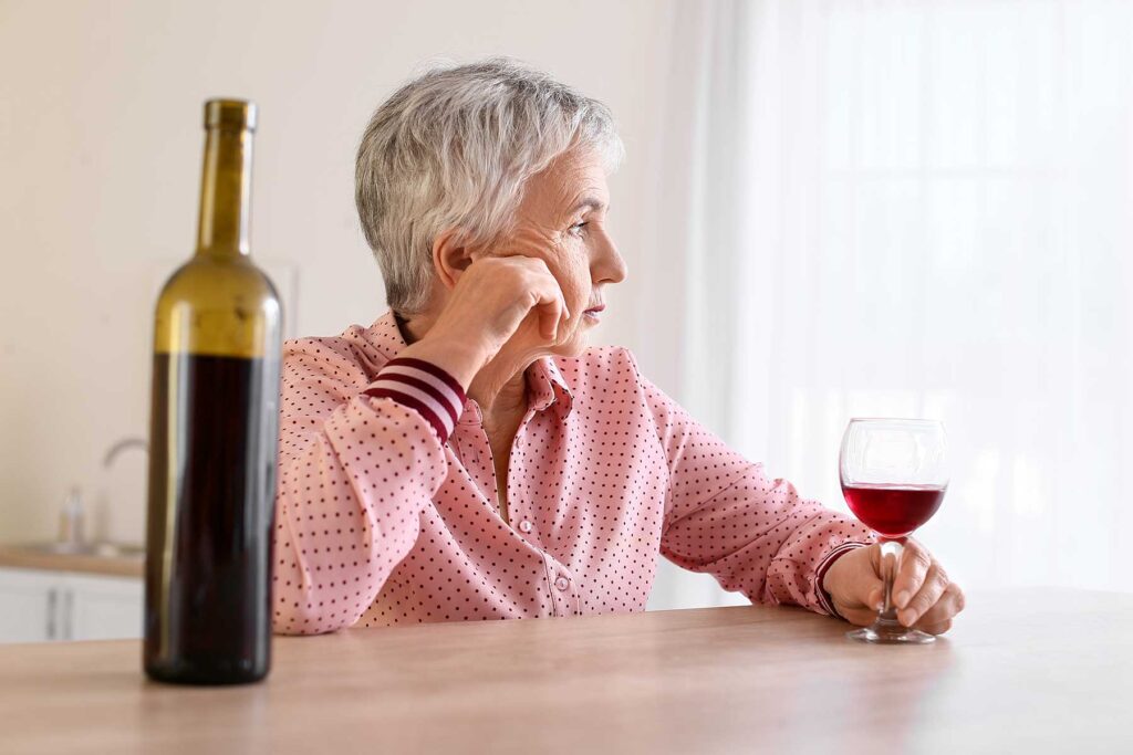 an older woman in a pini shirt sits at a table with a bottle of wine to show Alcohol and Breast Cancer